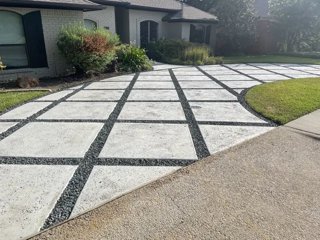 A front courtyard of large light gray concrete squares separated by dark gravel joints, curving past a lawn to a white brick house