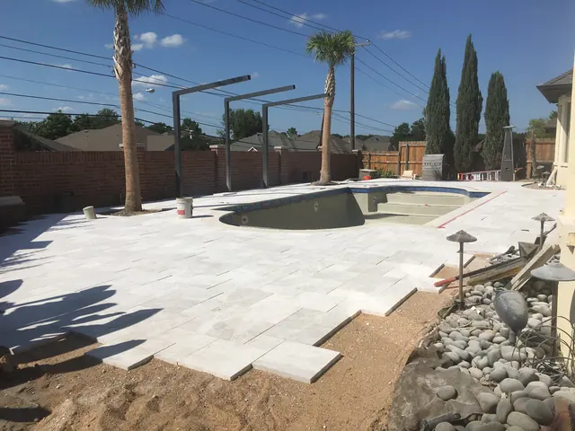 A pool deck part way through: pale large-format pavers laid out from the pool edge with bare bedding where the next courses go, and a river rock bed along the boundary