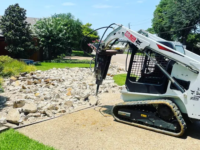 A tracked skid steer with a hydraulic breaker on the arm, standing on an exposed aggregate driveway beside the slab it has just broken into rubble