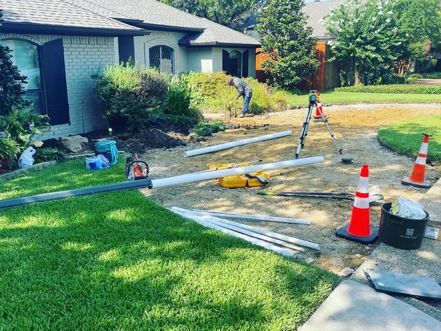 A front yard mid-job: laid sod on one side, graded ground on the other, lengths of pipe on the ground and a rotary laser level on a tripod