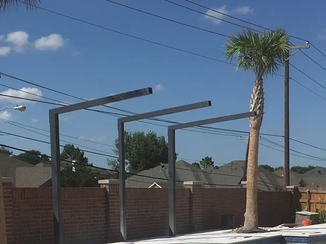 Three steel shade frames standing over a pool deck, square to each other and to the brick wall behind, with a palm and the neighboring roofs beyond