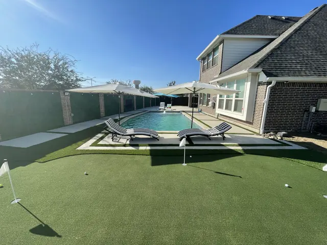 A concrete deck running around a pool between artificial turf, with a putting green in the foreground and a two-story brick house behind