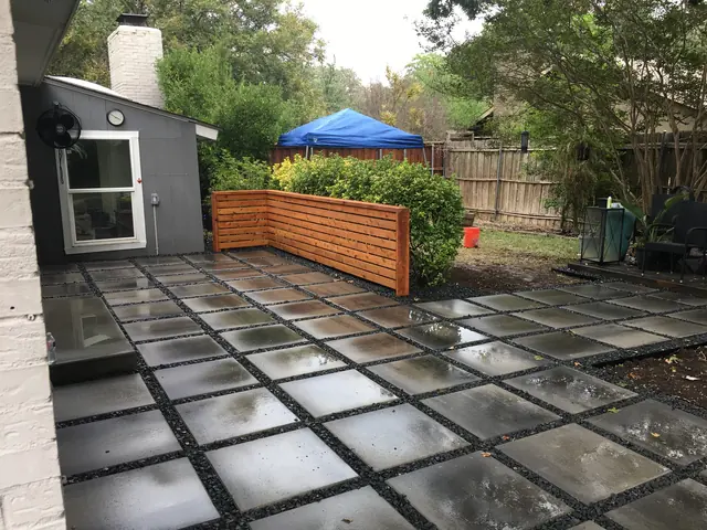 A patio of large gray units set in a grid with dark gravel joints, wet after rain, with a horizontal cedar screen along one side