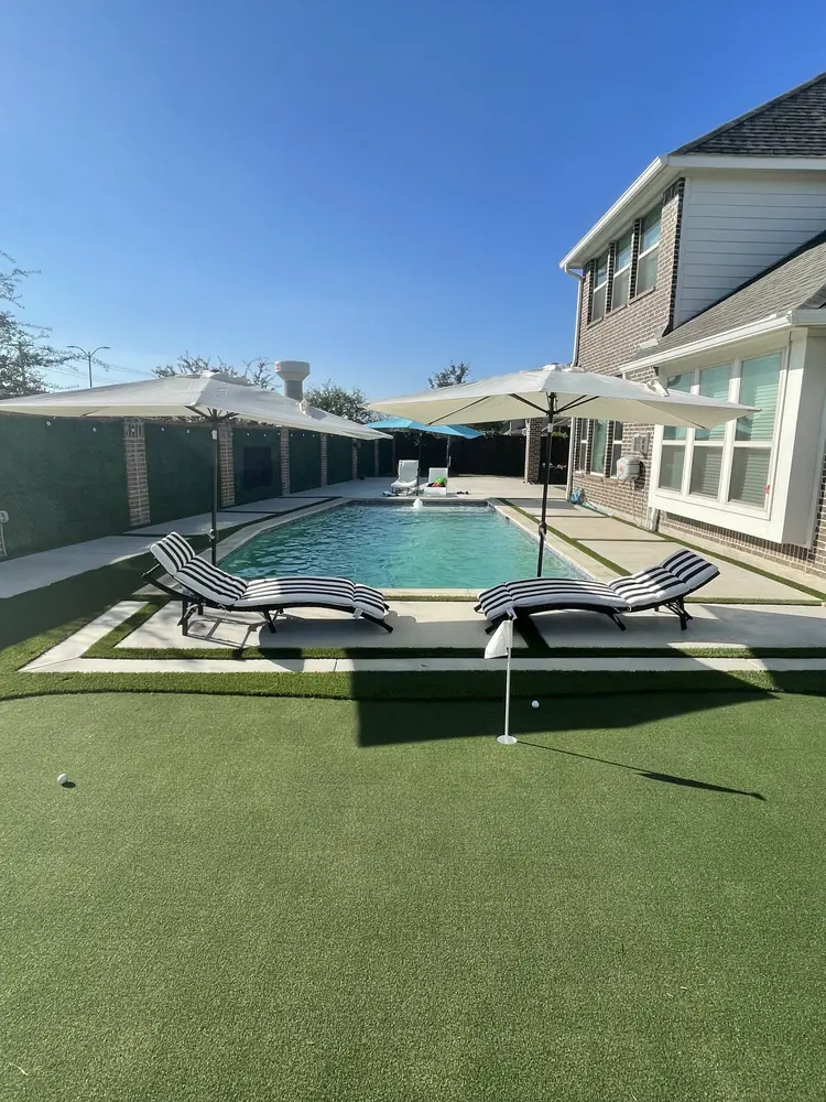 A finished back yard seen upright: a putting green in the foreground and a concrete deck running around a pool behind it
